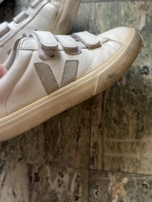Vita Veja sneakers med kardborreband - Snygga vita sneakers från Veja med tre kardborreband och grått V på sidan. Skorna är i läder med gummisula och har en klassisk låg profil som passar till det mesta. Perfekta för dig som gillar stilrena och trendiga sneakers de är lite smutsiga nu men behöver bara tvättas