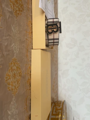 Rutig Burberry bälte med B-spänne - Snyggt rutig bälte från Burberry med klassiskt B-spänne i metall. Bältet har en beige bas med svarta, vita och röda ränder och är tillverkat i skinn. Perfekt accessoar för att lyfta din outfit.