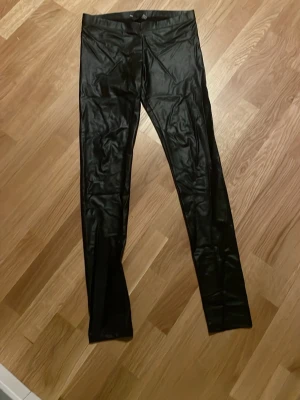 Svarta leggings i fuskskinn - Snygga svarta leggings i glansigt fuskskinn med hög midja och tight passform. Perfekta för dig som vill ha en edgy och trendig look. Materialet är stretchigt och följsamt för bästa komfort.