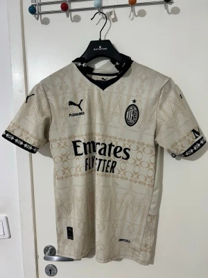 Beige AC Milan matchtröja Puma - Snygg beige AC Milan matchtröja från Puma med svarta detaljer och grafiskt mönster. Tröjan har korta ärmar, svart krage och loggor från Emirates, Fly Better och Pleasures. Tillverkad i funktionsmaterial Drycell som andas. Perfekt för fotbollsträning eller support.