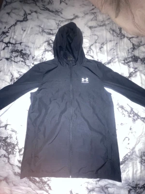 Svart vindjacka från Under Armour - Snygg svart vindjacka från Under Armour med huva och hel dragkedja framtill. Jackan har ett diskret logotryck på bröstet och är tillverkad i lätt, vindtätt material med meshfoder för extra komfort. Perfekt för blåsiga dagar och sportiga outfits.