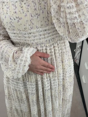 Lång blommig klänning med volangärm - Romantisk långklänning i ljus cremefärg med lila blommönster. Klänningen har volangiga, långa ärmar och smockdetaljer i midjan och överdelen. Tillverkad i ett lätt, plisserat material som ger en luftig känsla. Perfekt för sommarens alla tillfällen.