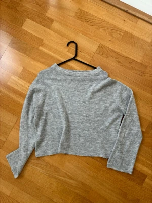 Ljusgrå croppad stickad tröja - Mysig ljusgrå stickad tröja med croppad passform och långa ärmar. Tröjan har en enkel, rak design och mjukt material som känns skönt mot huden. Perfekt för lager på lager och en avslappnad stil.