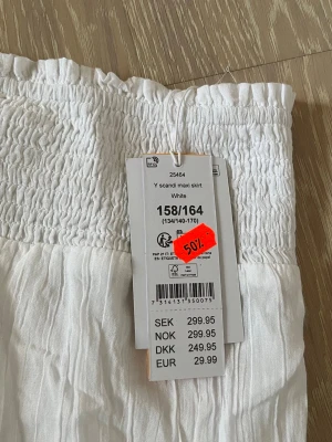 Vit långkjol med smock i midjan - Säljer en vit långkjol från gina med smockad midja och tunna volangdetaljer. Kjolen är luftig och har en bohemisk känsla, perfekt för varma dagar. Tillverkad i ett lätt och mjukt material som känns skönt mot huden.