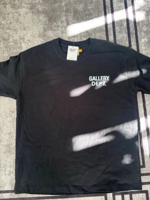 Svart t-shirt från Gallery Dept. - Snygg svart t-shirt från Gallery Dept. med vit logga tryckt på bröstet. Klassisk rund hals och korta ärmar. Tillverkad i mjuk bomull som känns skön mot huden. Perfekt för dig som gillar streetwear och enkel stil.