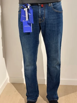Blå jeans från Jacob Cohen - Säljer ett par klassiska blå jeans från Jacob Cohen med raka ben och snygga kontrastsömmar. Jeansen har fem fickor, normal passform och en diskret logopatch bak i midjan. Tillverkade i mjukt bomullsmaterial för skön känsla hela dagen.