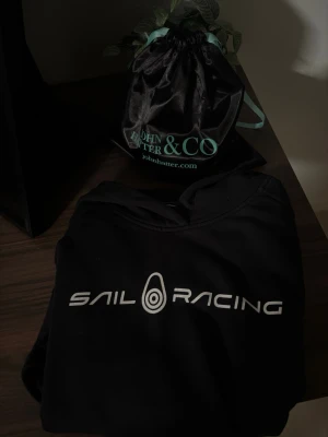 Svart hoodie från Sail Racing - Svart hoodie från Sail Racing med stor vit logga och text på bröstet. Klassisk huva och mjukt material som känns skönt mot huden. Perfekt för dig som gillar stilrena och sportiga plagg. Passar till jeans eller joggers för en avslappnad look.