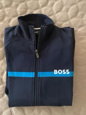 Marinblå ziptröja från BOSS - Snygg marinblå ziptröja från BOSS med blå rand och vit logga över bröstet. Tröjan har hög krage, hel dragkedja och två fickor framtill. Perfekt för en sportig och avslappnad stil.