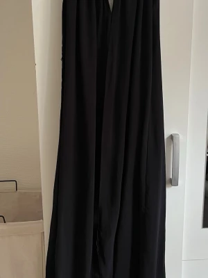 Svart långklänning med omlott - Säljer en svart långklänning med omlottdesign och ärmlös passform. Klänningen har en luftig och flowy siluett med en liten v-ringning framtill. Perfekt för dig som gillar stilrena och eleganta plagg.