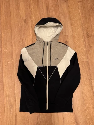 Svart, grå och vit hoodie från BOSS  - Snygg hoodie från BOSS i svart, grått och vitt med dragkedja framtill och huva. Stora färgblock ger en modern look och BOSS-loggan är broderad på bröstet. Tillverkad i mjukt bomullsmaterial som känns skönt mot huden. Perfekt för en avslappnad streetwear-stil. 