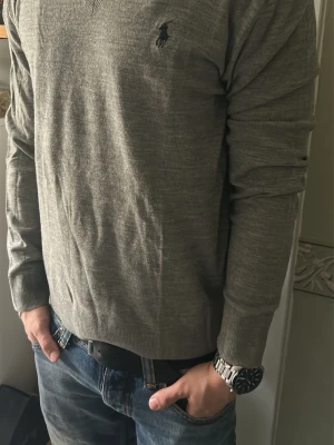 Grå stickad tröja Polo Ralph Lauren - Snygg grå stickad tröja från Polo Ralph Lauren med diskret logga broderad på bröstet. Tröjan har v-ringning, långa ärmar och är tillverkad i en mjuk ull- och polyestermix. Perfekt för lager på lager och enkel att matcha med jeans.