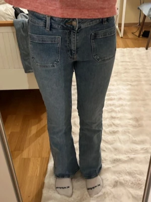 Blå bootcut jeans med stora fickor - Säljer ett par ljusblå bootcut jeans med stora fyrkantiga fickor fram och bak. Jeansen har hög midja och klassisk knappgylf. Tillverkade i denim med en snygg tvätt som ger en retrovibe. Perfekta för dig som gillar en avslappnad men trendig stil.