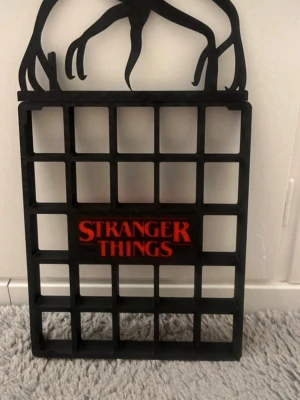 Stranger Things hylla svart - Säljer en svart hylla med Stranger Things-logga i rött och en siluett av en Demogorgon på toppen. Hyllan har flera fack och är perfekt för småprylar eller samlarobjekt. Tillverkad i plast och har en cool, unik design för fans av serien.