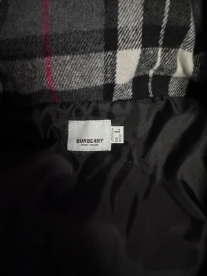Svart pufferjacka från Burberry - Säljer en svart pufferjacka från Burberry med klassiskt rutigt foder i svart, vitt och rött. Jackan har en modern, puffig design och är tillverkad i slitstarkt material med mjukt innerfoder. 