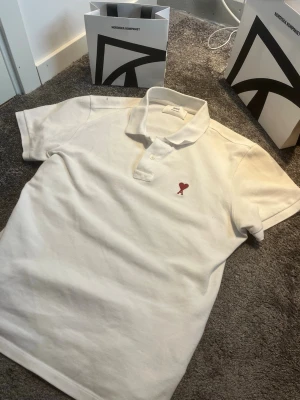Vit pikétröja från Ami Paris - Vit pikétröja från Ami paris i mycket bra skick. Saknar defekter och är en väldigt stilren piké som passar bra till jeans. Hör av er vid frågor!