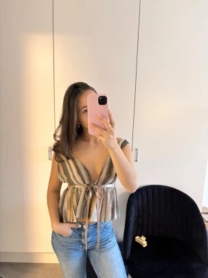 Randig topp med knytband - Säljer en snygg randig topp i beige, brun och vit med djup v-ringning och breda axelband. Toppen har ett knytband i midjan och volang nedtill. Perfekt till jeans eller kjol för en trendig look.
