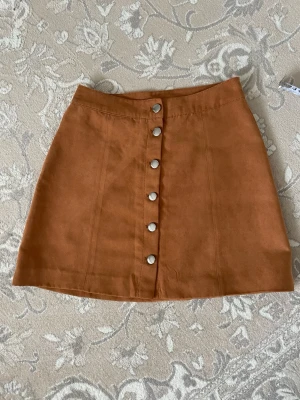 Brun kortkjol med knappar - Snygg brun kortkjol med silverfärgade knappar framtill. Kjolen har en A-linjeform och är tillverkad i ett mjukt material som liknar mocka. . Knappar man kan knäppa upp