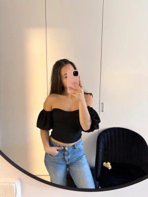 Svart offshoulder topp med puffärm - Trendig svart offshoulder topp med puffiga korta ärmar och figurnära passform. Toppen är croppad och har en snygg, enkel design som passar perfekt till jeans eller kjol. Tillverkad i mjukt material för en bekväm känsla.