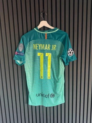 Barcelona Neymar Jr matchtröja - Snygg Barcelona matchtröja från Nike med Neymar Jr och nummer 11 på ryggen. Tröjan är turkos med tunna vita ränder, officiella Champions League-patchar på ärmarna och klubbmärke samt Qatar Airways-logga på bröstet. Tillverkad i lätt och ventilerande material.