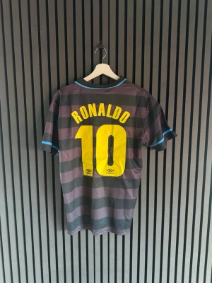 Inter Milan Ronaldo tröja Umbro - Säljer en svart och grå randig Inter Milan fotbollströja från Umbro med blå detaljer. Tröjan har nummer 10 och Ronaldo på ryggen, samt Pirelli-logga och klubbmärke på bröstet. Perfekt för dig som gillar klassiska fotbollströjor.