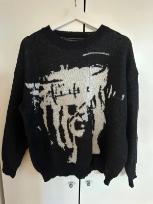 Svart stickad tröja med ansiktsmotiv - Cool svart stickad tröja med ett stort grafiskt ansiktsmotiv i vitt på framsidan. Tröjan har rund halsringning, långa ärmar och ribbade muddar vid ärmslut och nederkant. Oversized passform för en avslappnad look. 