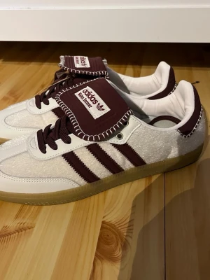 Adidas Wales Bonner vita sneakers - Unika Adidas Wales Bonner sneakers i vitt och vinrött med klassiska tre ränder på sidan. Skorna har en låg silhuett, grov sula i gummi och detaljer i mocka och läder. Tungan har kontrastsömmar och Wales Bonner-logga. Perfekta för dig som gillar retrostil och coola detaljer.