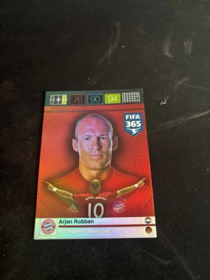 FIFA 365 samlarkort Arjen Robben - Samlarkort från Panini FIFA 365 med Arjen Robben i Bayern München. Kortet har en röd och svart design med klubbmärke och spelarstatistik. Baksidan visar en siluett av en fotbollsspelare och en aktiveringskod för onlineutmaning.