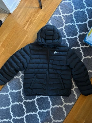 Svart pufferjacka från Nike - Säljer en svart pufferjacka från Nike med huva och dragkedja framtill. Jackan har två sidofickor och den klassiska Nike-loggan i vitt på bröstet. Perfekt för kalla dagar och riktigt skön att bära tack vare det vadderade materialet.