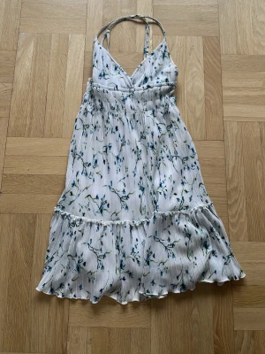 Vit blommig långklänning med volang - Söt långklänning i vitt med blått blommönster och tunna axelband. Klänningen har v-ringning, smock i midjan och volang nertill för en luftig känsla. Perfekt för varma dagar och somriga tillfällen. Banden har en liten missfärgning 