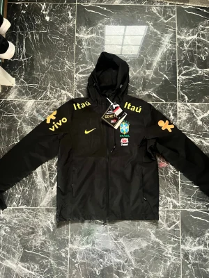Svart Brasil Nike vindjacka med huva - Snygg svart vindjacka från Nike med Brasilien CBF-logga på bröstet och gula sponsortryck på ärmar och axlar. Jackan har huva, dragkedja framtill och är tillverkad i slitstarkt GORE-TEX-material. Perfekt för dig som gillar fotboll och streetwear.
