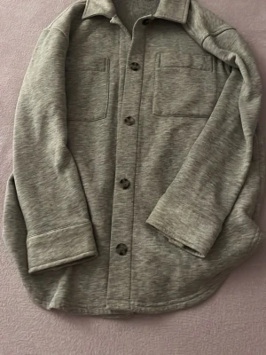 Grå overshirt med fickor - Stilren grå overshirt med två stora bröstfickor och knäppning framtill. Overshirten har en klassisk krage och långa ärmar med breda muddar. Materialet ser mjukt och lite stretchigt ut, perfekt för en avslappnad look.