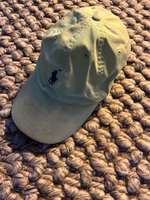 Ljusgrön keps från Polo Ralph Lauren - Snygg ljusgrön keps från Polo Ralph Lauren med klassisk böjd skärm och mörkblå broderad logga framtill. Kepsen har ventilerande öljetter och justerbar rem bak. Tillverkad i mjuk bomull som ger en avslappnad och stilren look.