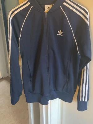Marinblå Adidas hoodie med dragkedja - Snygg marinblå hoodie från Adidas med klassiska vita ränder längs ärmarna och Adidas-logga på bröstet. Dragkedja framtill, två fickor och ribbade muddar. Tillverkad i mjukt och bekvämt material, perfekt för en sportig och avslappnad stil.