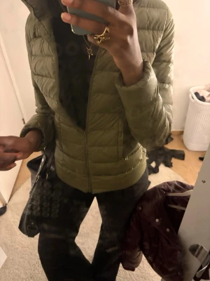 Olivgrön dunjacka med hög krage - Säljer en olivgrön dunjacka med hög krage och quiltad design. Jackan har dragkedja framtill och två sidofickor. Perfekt för vindiga dagar och har en stilren, enkel look som passar till det mesta.
