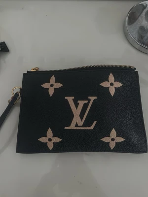 Svart Louis Vuitton kuvertväska - Snygg svart kuvertväska från Louis Vuitton med klassiska monogramdetaljer i beige och guldfärgad dragkedja. Väskan har en avtagbar handledsrem och röd insida. Köpt på Louis Vuitton butiken tillsammans med en till väska så kvitto medföljer!💕