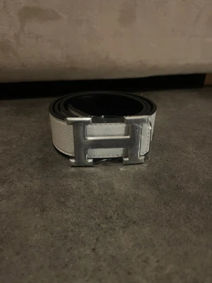 Vitt skärp med H-spänne - Snyggt vitt skärp  med klassiskt stort H-spänne i silverfärgad metall. Bältet är gjort i skinn och har en stilren, modern look som passar till många outfits. Perfekt accessoar för dig som vill ha något exklusivt och trendigt.
