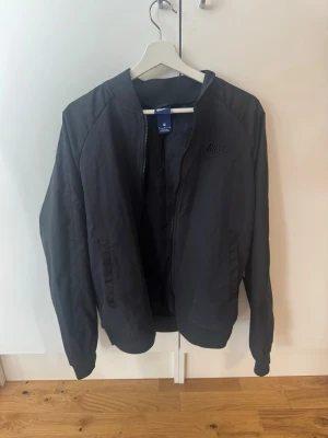 Svart bomberjacka från Nike - Snygg svart tunn bomberjacka från Nike med diskret logga på bröstet. Jackan har dragkedja framtill, ribbade muddar vid ärmslut och nederkant samt två fickor med dragkedja. Klassisk bombermodell i ett lätt material som passar perfekt till streetwear.