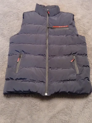 Mörkblå dunväst med dragkedjor - Snygg mörkblå dunväst med hög krage och tre fickor med dragkedja, varav en bröstficka med röd detalj. Västen har quiltad design och stängs med dragkedja framtill. Perfekt för lager-på-lager och håller dig varm under kyliga dagar.