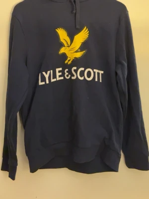 Marinblå hoodie från Lyle & Scott - Snygg marinblå hoodie från Lyle & Scott med stor gul örn och vit logga på bröstet. Klassisk huva med dragsko och långa ärmar. Perfekt för en avslappnad och sportig stil.