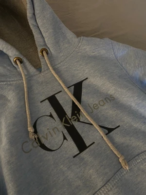 Ljusblå hoodie från Calvin Klein - Snygg ljusblå hoodie från Calvin Klein Jeans med stor CK-logga på bröstet. Hoodien har huva med dragsko, magficka och ribbade muddar. Mjuk insida och klassisk passform, perfekt för en avslappnad och trendig look. Passar till 9-11 år ☺️