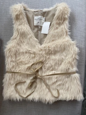 Ny fuskpälsväst från H&M i str.S - Säljer en fluffig krämvit fuskpälsväst från H&M med mjukt foder och ett smalt beige knytband i midjan. Helt ny och oanvänd med prislappen kvar. Storlek:S