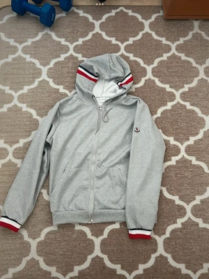 Grå Moncler hoodie med dragkedja - Snygg grå hoodie från Moncler med dragkedja framtill och huva. Röd, vit och marinblå rand på huva och ärmslut ger en sportig vibe. Moncler-logga på vänster ärm. Perfekt för en chill och stilren look.