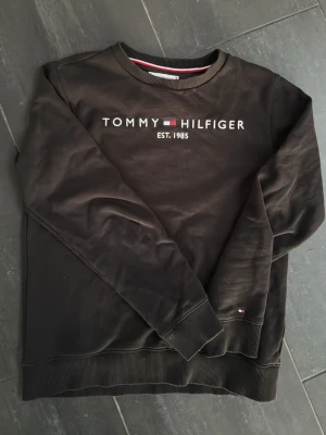 Svart sweatshirt Tommy Hilfiger - Svart sweatshirt från Tommy Hilfiger med broderad logga och text 'EST. 1985' på bröstet. Tröjan har rund halsringning, ribbade muddar och en liten flaggdetalj vid ärmslutet. Perfekt för en avslappnad och stilren look.