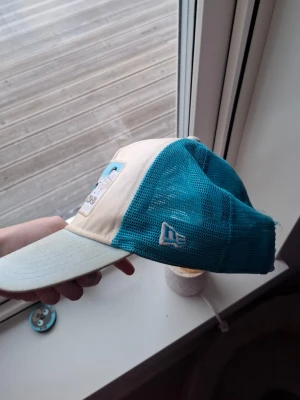 Beige och turkos Mykonos keps - Säljer en beige och turkos keps från New Era med mesh på sidorna och broderad Mykonos-patch framtill. Framsidan har motiv av hav, sol och vita hus. Skärmen är böjd och kepsen har klassisk trucker-modell. Perfekt för dig som gillar unik stil och semester-vibes.