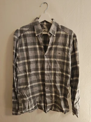 Gråvit rutig skjorta från Lacoste - Klassisk grå och vit rutig skjorta från Lacoste med button-down krage och diskret logga på bröstet. Skjortan har långa ärmar och knäppning framtill. Perfekt för en avslappnad men ändå stilren look.