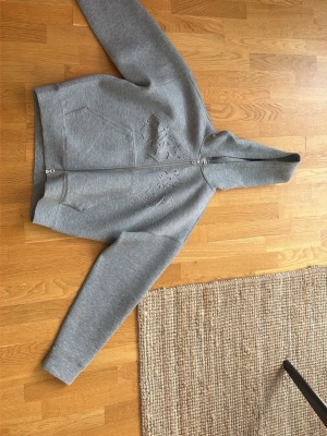 Grå hoodie med dragkedja från Weekday - Säljer en grå hoodie från Weekday med dragkedja framtill och ett stort, ton-i-ton broderat tiger-motiv på bröstet. Hoodien har huva och två fickor framtill. Tillverkad i mjuk bomullsblandning som känns riktigt skön att ha på sig.