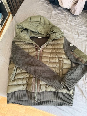 Olivgrön moncler jacka med stickade detaljer - Snygg olivgrön dunväst med stickade mörkgröna ärmar och muddar. Västen har en stor huva, två fickor med dragkedja och en extra ficka på ärmen. Perfekt för lager på lager och streetwear-vibe. Storlek L passar även M