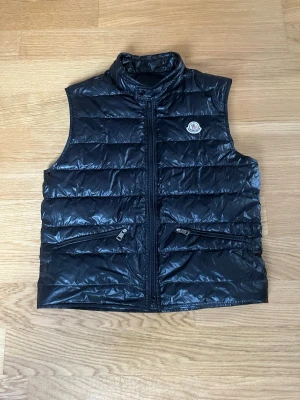 Svart dunväst från Moncler - Snygg svart dunväst från Moncler med hög krage och två dragkedjeförsedda fickor framtill. Västen har ett quiltat mönster och Moncler-logga på bröstet. Perfekt för lager-på-lager och ger en clean look.