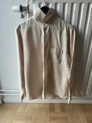 Beige skjorta; A Day’s March - Stilren beige skjorta med hög krage och bröstficka. Skjortan har lång ärm och knäppning framtill. Perfekt för dig som gillar minimalistisk och clean stil.