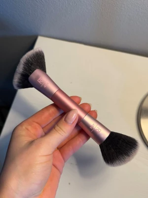 Rosa dubbeländad sminkborste från Real Techniques - Säljer en rosa dubbeländad sminkborste från Real Techniques. Ena sidan har en snedställd borste och andra sidan en rundad, fluffig borste. Handtaget är i metall och borsten är gjorda av mjuka syntetfibrer. Perfekt för contouring och puder💗